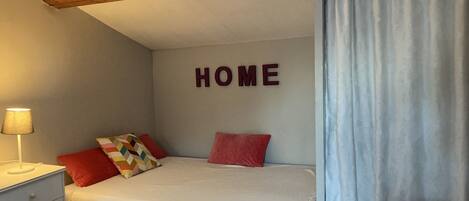 A bedroom with a bed, a nightstand, a lamp, a window with a view, and a 'HOME' sign on the wall.