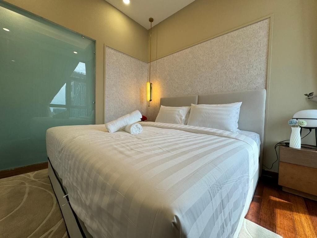 A neatly made bed with white linens, a bedside table with a lamp, and a view of a building through a glass door.
