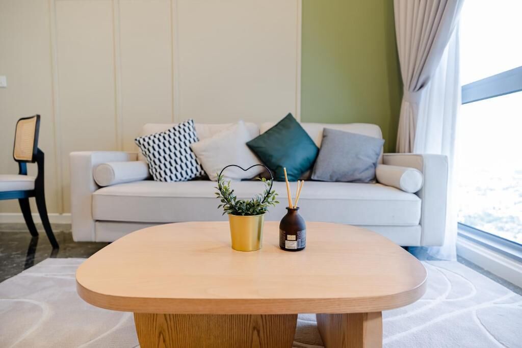 Ein modernes Wohnzimmer mit einem Holztisch, einer Topfpflanze und einem Kerzenhalter. Ein weißes Sofa mit gemusterten Kissen befindet sich im Hintergrund.