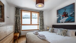 A bedroom with a large bed, a wooden dresser, a bench, a window with curtains, a painting, and a lamp.