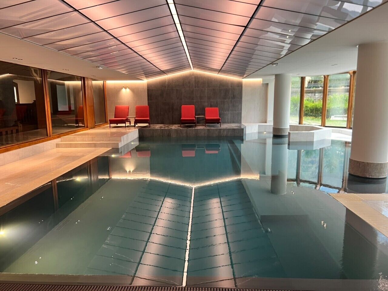 Ein moderner, überdachter Hallenpool mit roten Liegestühlen und Blick auf Grünflächen.