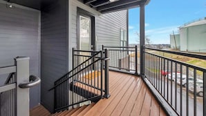 A covered outdoor area with a wooden deck, black metal railing, and a door leading to a building with gray siding. - Long Term Condo with Pool, Gym, Balcony, Parking   |   Sleeps 6, King Suite + Sleeper Sofa,  Minutes to Downtown & Five Points, Perfect for Long Stays and Remote Work,  24 Hour Hospitality (Nashville)