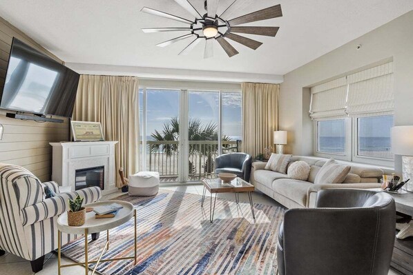 A spacious living room with a fireplace, a ceiling fan, a large window with a view, and a television mounted on the wall. - Celadon 109 - Free Seasonal Beach Chairs! (Panama City Beach)