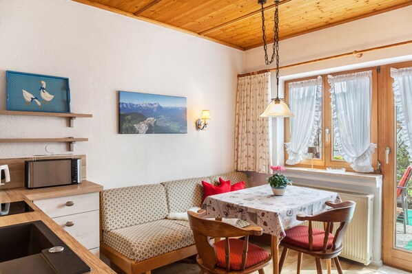 A cozy room with a wooden ceiling, a dining table with chairs, a sofa, a microwave, a small shelf, and a window with curtains.
