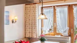 A cozy room with a dining table, a sofa with red cushions, a wall-mounted lamp, and a window with curtains.