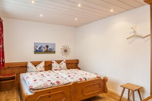 A wooden bed with a floral bedspread, a small bedside table, a wall-mounted lamp, and a picture of mountains.