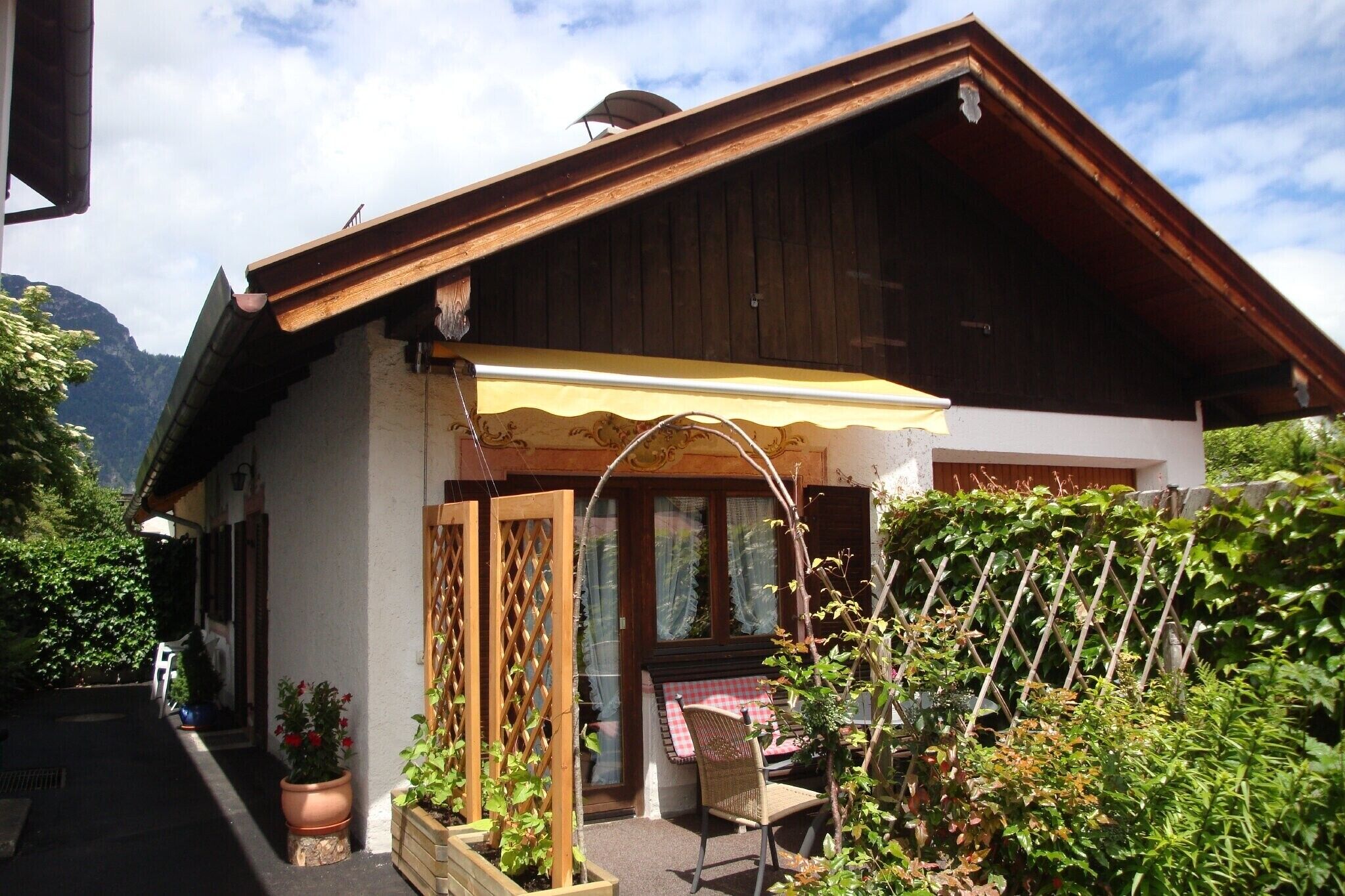 A chalet with a wooden exterior, a covered porch, and a garden area with potted plants and a chair.