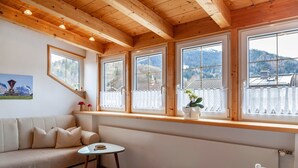 A cozy room with a wooden ceiling, large windows with white curtains, a sofa, a small table, and a view of mountains. - Apartment 'Wank' with Mountain View, Balcony and Wi-Fi (Garmisch-Partenkirchen)