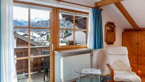 A room with a large window, a chair, a table, and a wooden cabinet. - Apartment 'Wank' with Mountain View, Balcony and Wi-Fi (Garmisch-Partenkirchen)