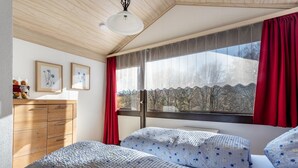 Ein Schlafzimmer mit einem Holzkapständer, zwei Betten mit Blumenmuster-Bettwäsche, einem Fenster mit roten Vorhängen und floralem Bild an der Wand.