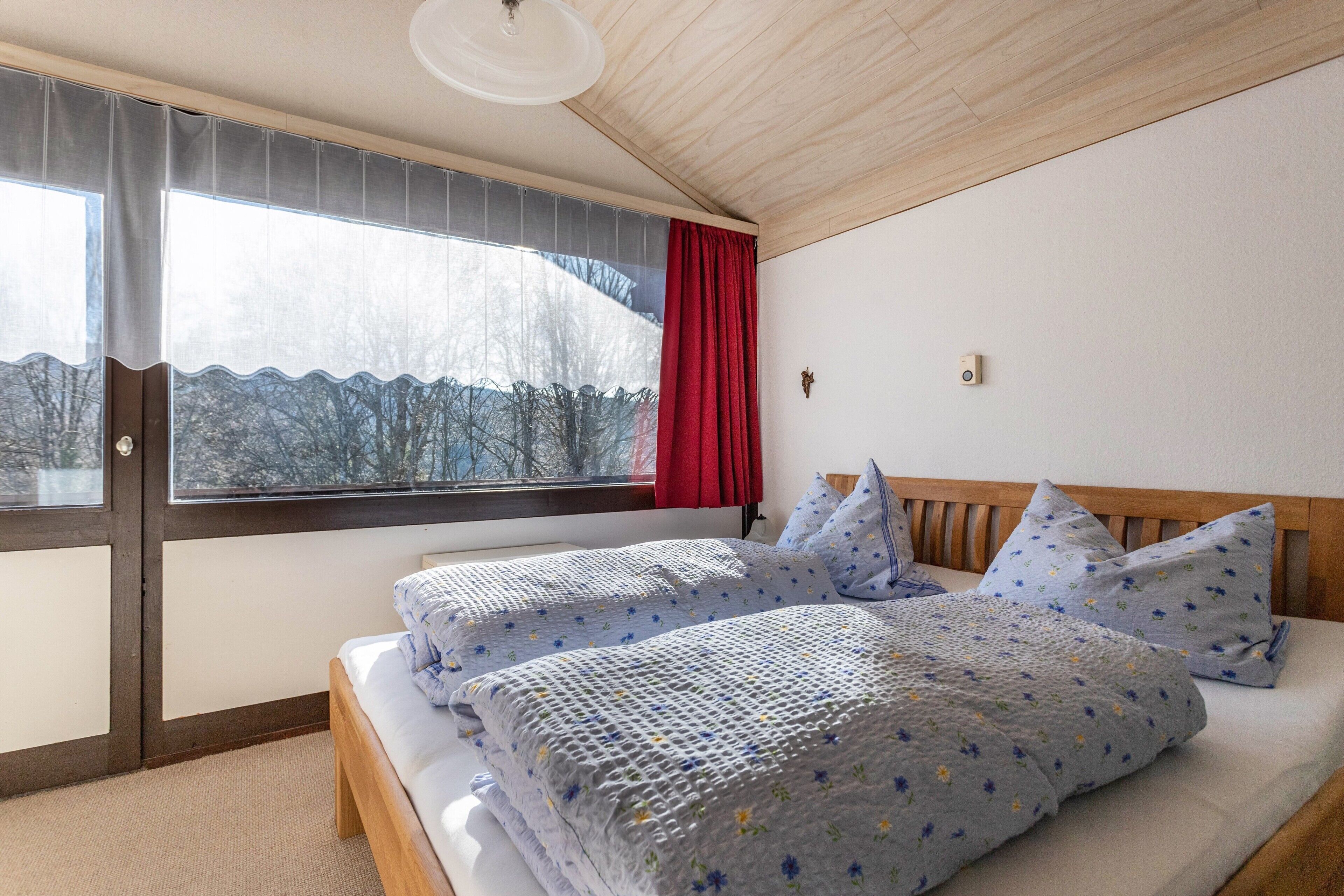 Ein Schlafzimmer mit großem Fenster, einem Bett mit floraler Bettwäsche und einer Holzbalkendecke.