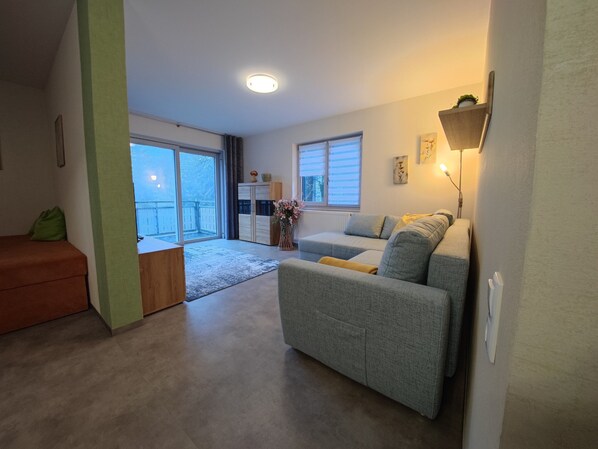 A living room with a sofa, a coffee table, a sliding glass door leading to a balcony, and a wall-mounted lamp.