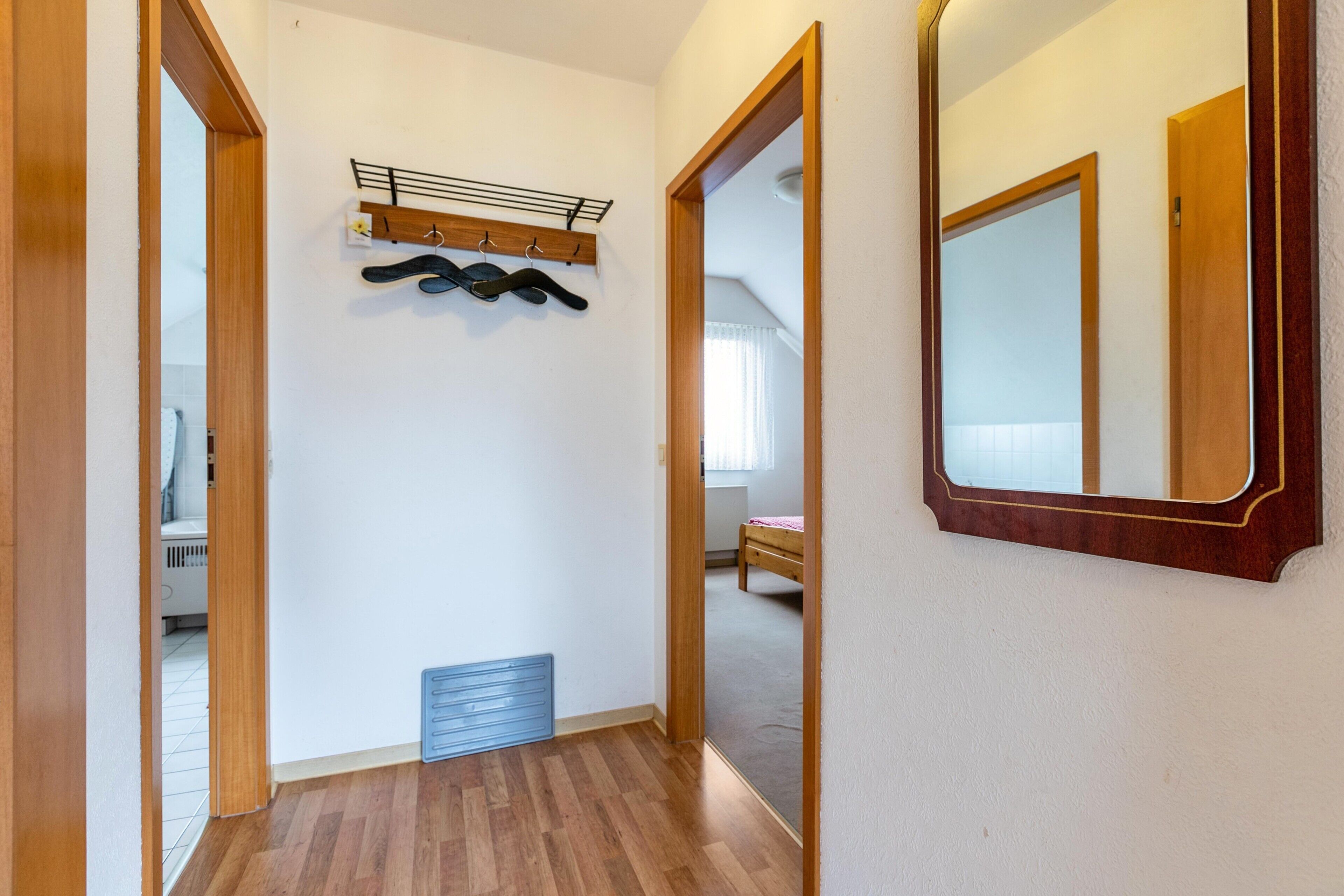 A hallway with wooden flooring, a coat rack, a vent, and a mirror on the wall.