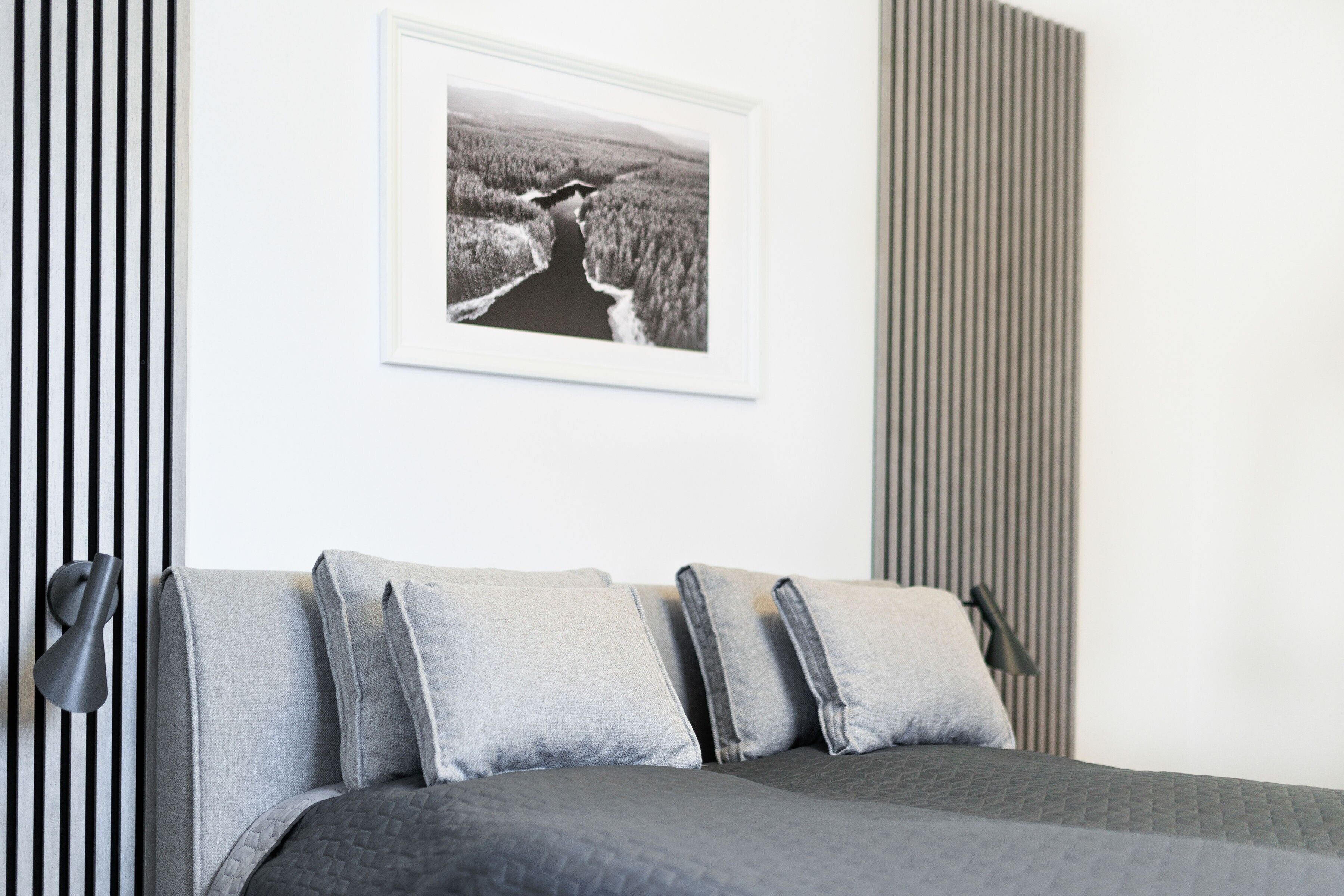 A bed with grey bedding, a framed landscape photo above it, and a striped wall to the left.