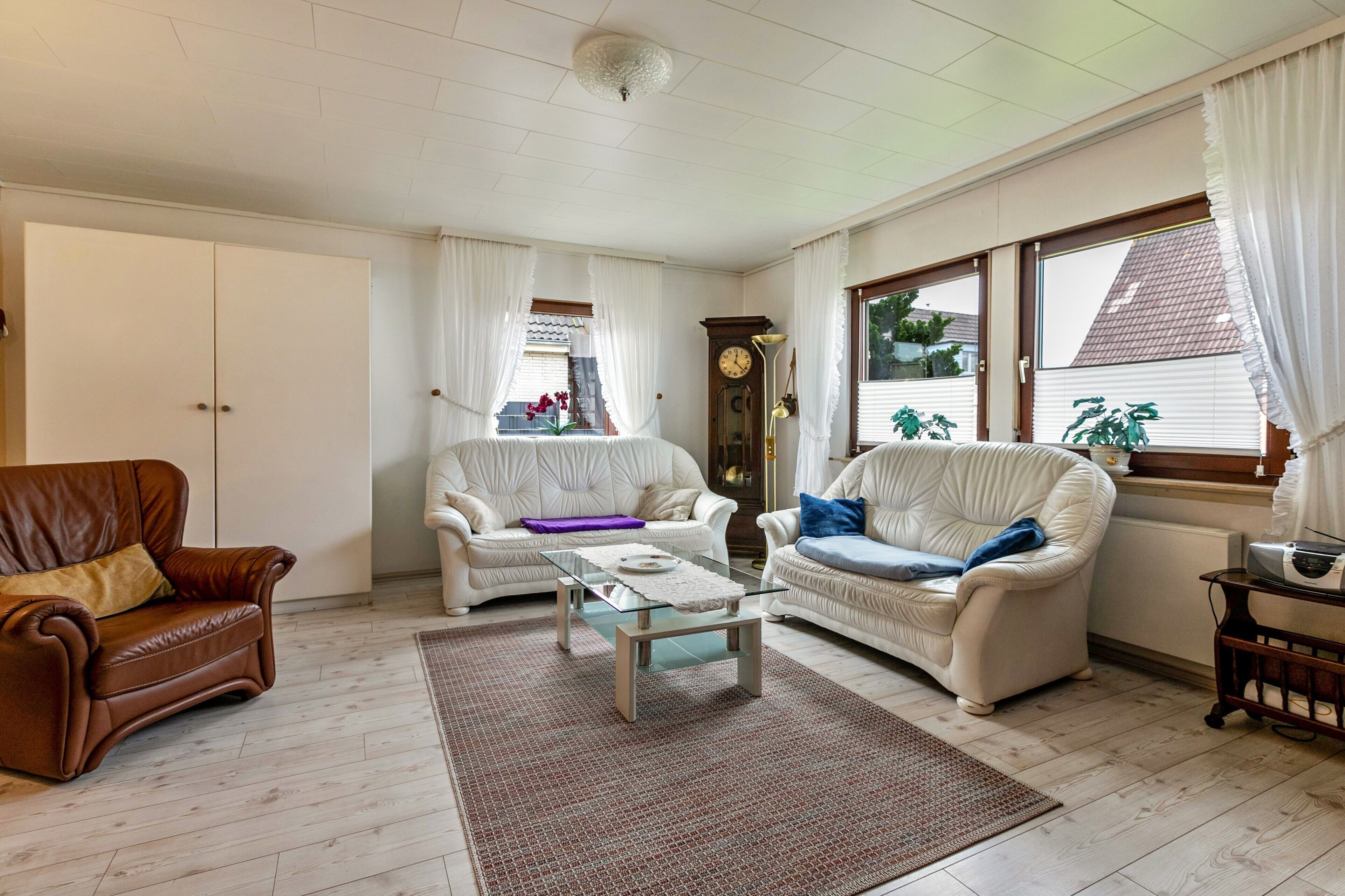 Un salon avec un fauteuil en cuir marron, deux canapés blancs, une table basse en verre, un tapis à motifs, une horloge et une fenêtre avec des rideaux.