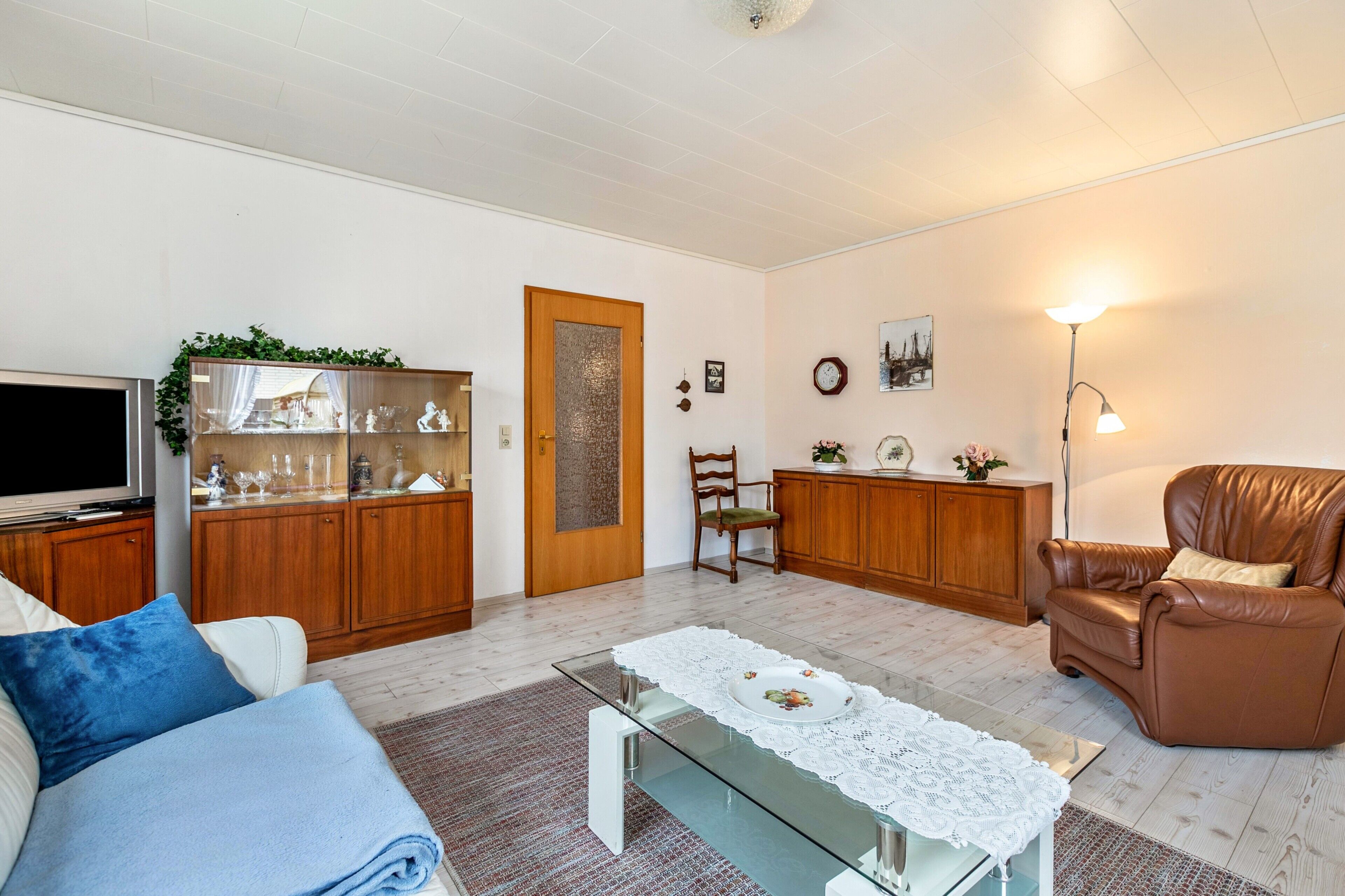 A living room with a sofa, a glass coffee table, a brown leather armchair, a wooden cabinet, a television, and a door with a decorative panel.