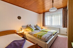 Een slaapkamer met een houten plafond, een bed met een wit matras, een nachtkastje met een lamp en een raam met gordijnen.