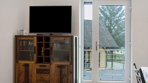 Une pièce avec un téléviseur posé sur un meuble en bois, une porte-fenêtre coulissante donnant sur l’extérieur et un coin repas avec une table et des chaises.