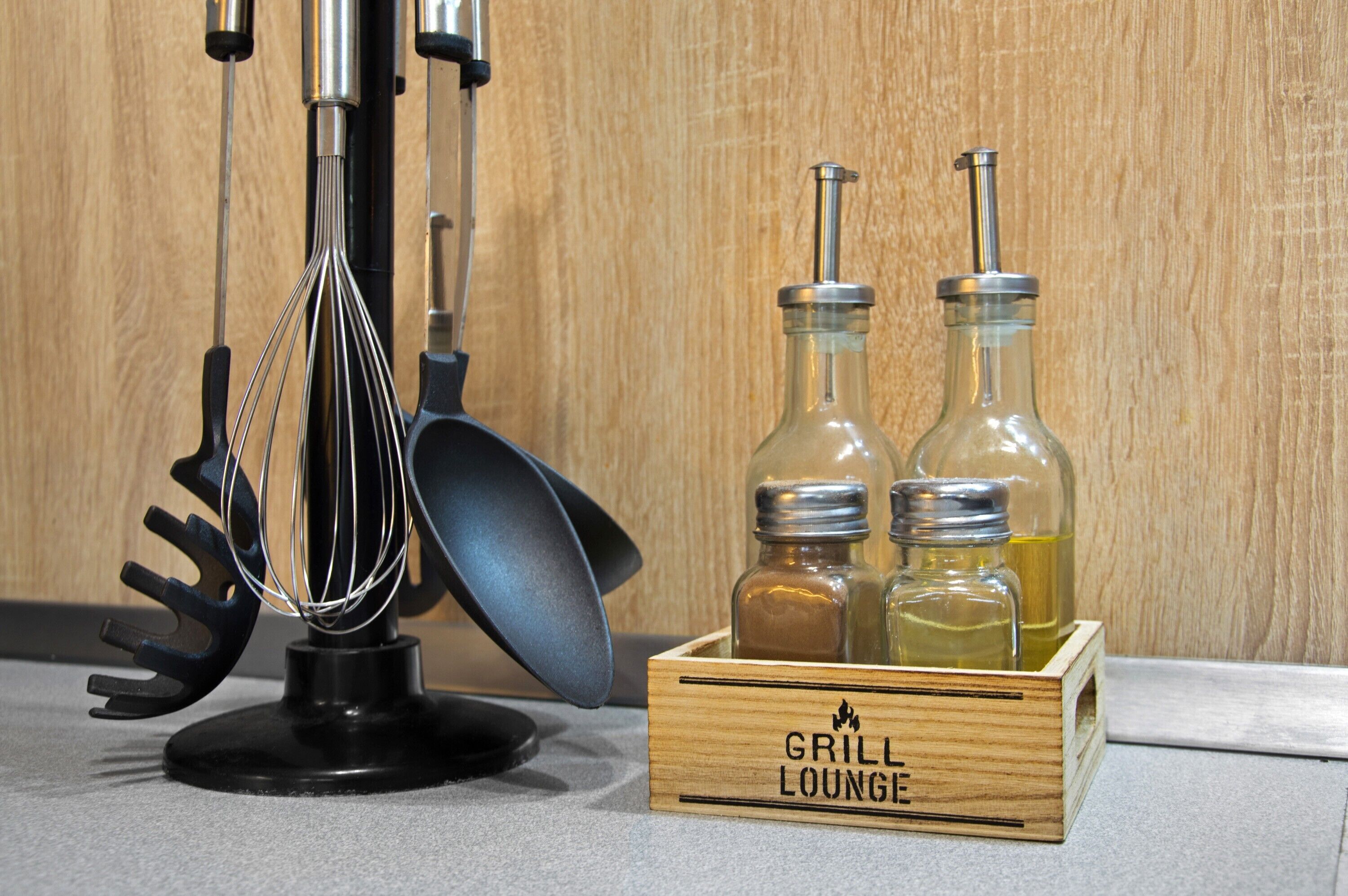 Un coin cuisine comprenant un fouet, une spatule et une boîte en bois portant l’inscription « GRILL LOUNGE » et contenant deux bouteilles.