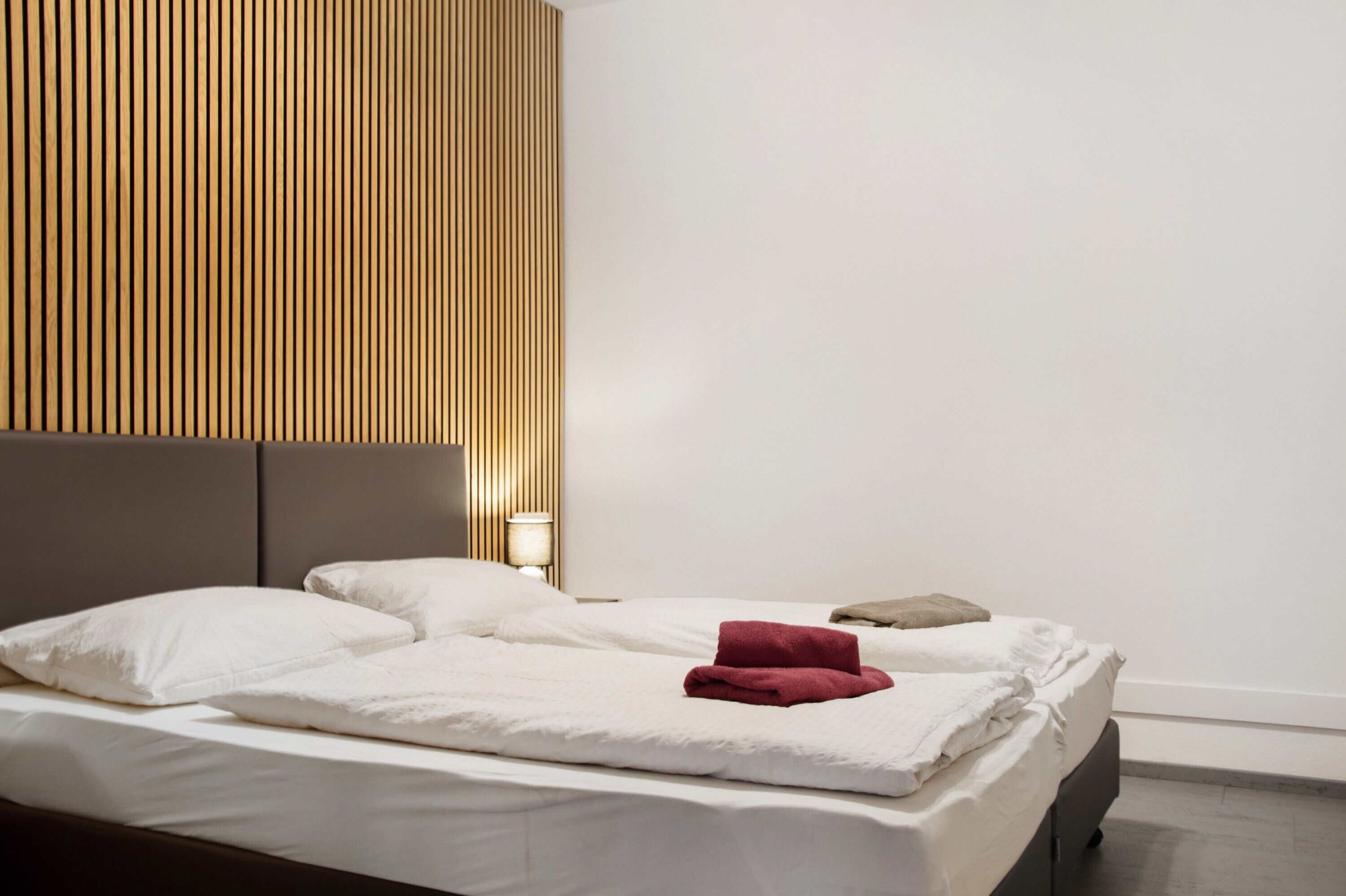 A neatly made bed with white linens and a red towel, a bedside lamp, and a wooden slat headboard.