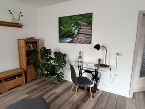 Un salon avec un plancher en bois, une chaise noire, une table en verre, une bibliothèque, une plante en pot, un tableau représentant un sentier forestier et une lampe.
