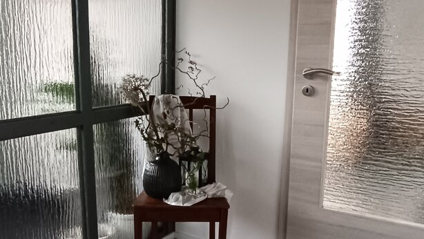 A room with a wooden floor, a chair with a vase of flowers, a large window with frosted glass, and a door with a frosted glass panel. - Apartment 'Papperitz' with Mountain View, Private Terrace and Wi-Fi (Uhlstädt-Kirchhasel)