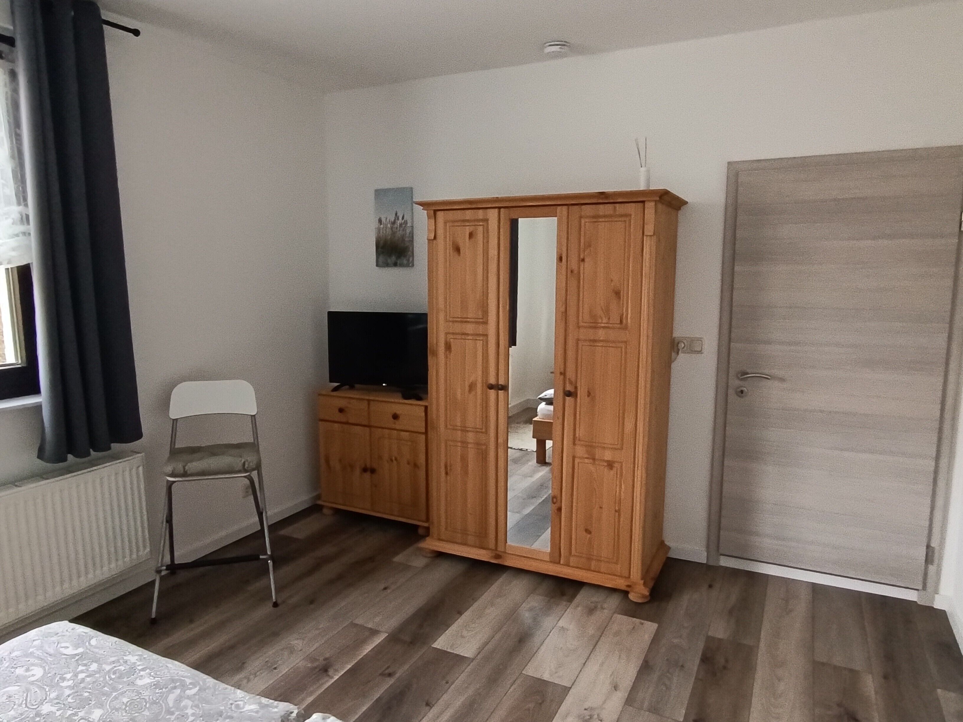 A room with a wooden wardrobe, a television on a cabinet, a chair, and a radiator.