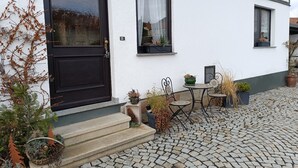 A white building with a black door, a window with a curtain, and a small outdoor seating area with chairs and a table. - Apartment 'Papperitz' with Mountain View, Private Terrace and Wi-Fi (Uhlstädt-Kirchhasel)