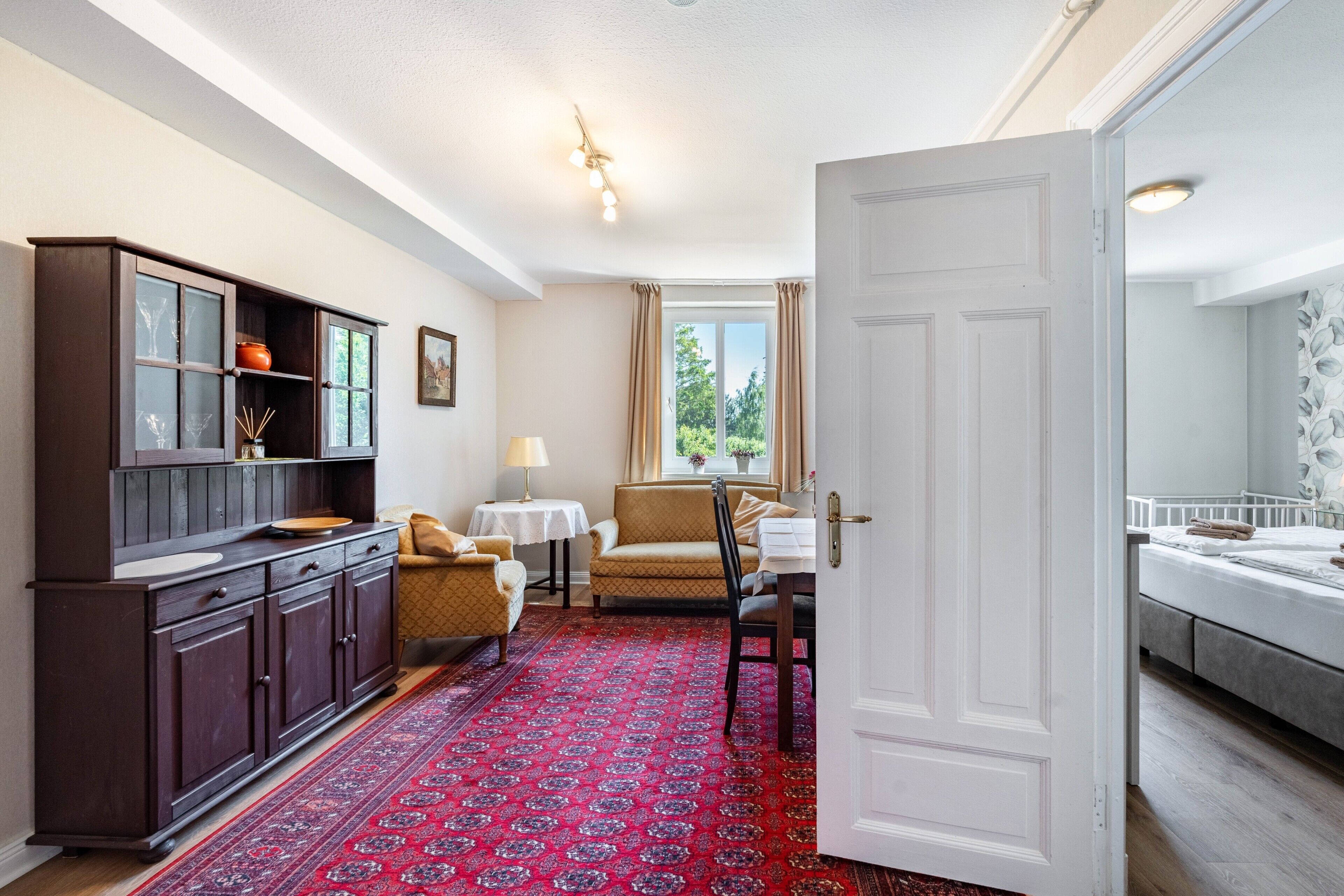 Una habitación con un armario de madera oscura, un tapete rojo estampado, un sofá color beige, una mesita con una lámpara y una cama con cabecera blanca.