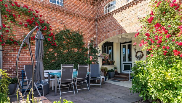 A brick building with a patio area featuring a table and chairs, a hanging chair, and potted plants. - Apartment 'Ferienwohnung Bauernhof Rohde' with Shared Terrace, Private Garden and Wi-Fi (Gremersdorf)