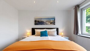 Une chambre à coucher avec un lit, un tableau représentant une plage, deux lampes de chevet et une fenêtre avec des rideaux.