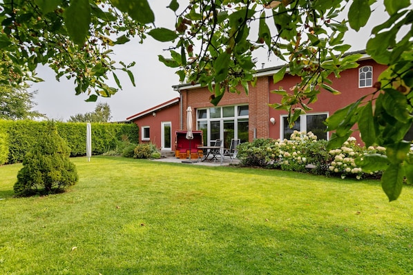 Ein rotes Ziegelhaus mit gepflegtem Rasen, einer Terrasse mit Gartenmöbeln und einem Hecken im Vordergrund.