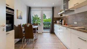 Une cuisine moderne avec des meubles blancs, un coin repas avec des chaises marron et une vue à travers la porte vitrée.
