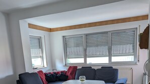 A living room with a sectional sofa, a round ottoman, a small table, and large windows with blinds.