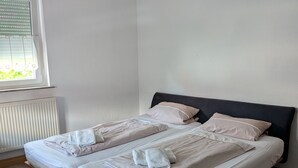 A bedroom with two beds, a window with blinds, a radiator, and wooden flooring.