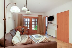 A living room with a sofa, a dining table with chairs, a TV mounted on the wall, and a wooden door.