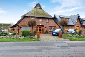 Un bâtiment traditionnel en briques, avec un toit de chaume, un chemin de pierre et une voiture garée devant.