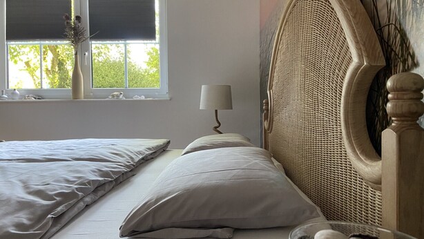 Un dormitorio con cama, almohadas, una lámpara de noche, una ventana con persianas y un adorno en la pared.