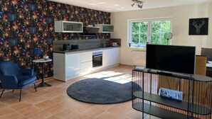 Una cocina moderna con una isla central, un comedor con una silla azul, un televisor sobre un mueble bajo y un ventanal con vistas a zonas verdes.