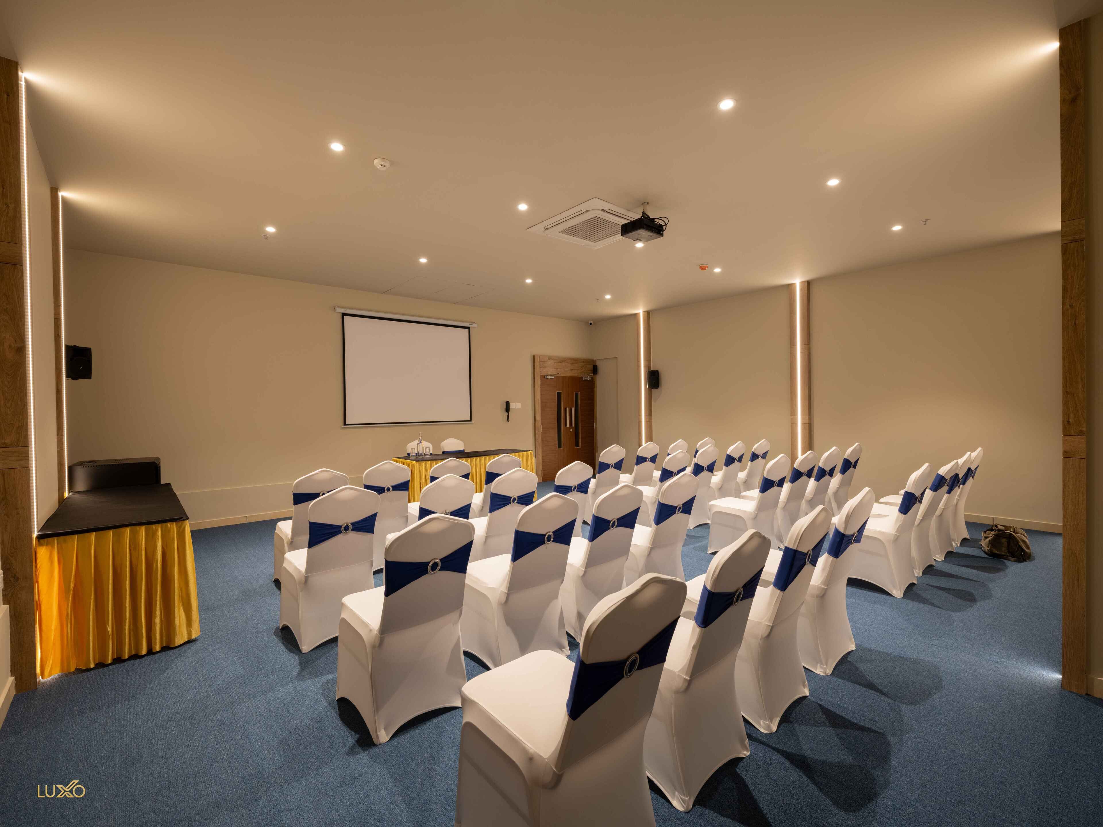 A conference room with rows of white chairs with blue sashes, a podium, a projector screen, and a carpeted floor.