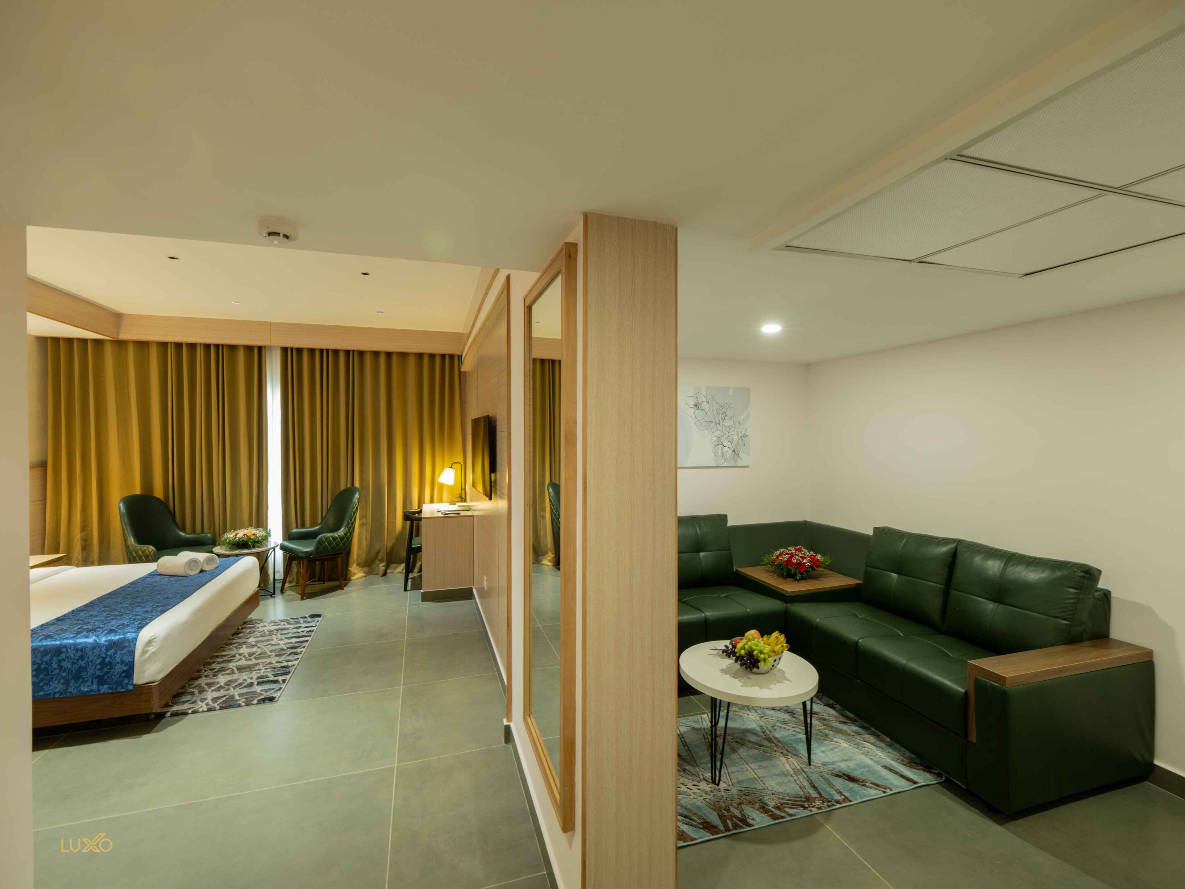 A hotel room with a bed, two chairs, a sofa, a small table with fruit, and a painting on the wall.