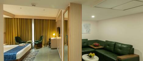 A hotel room with a bed, two chairs, a sofa, a small table with fruit, and a painting on the wall.