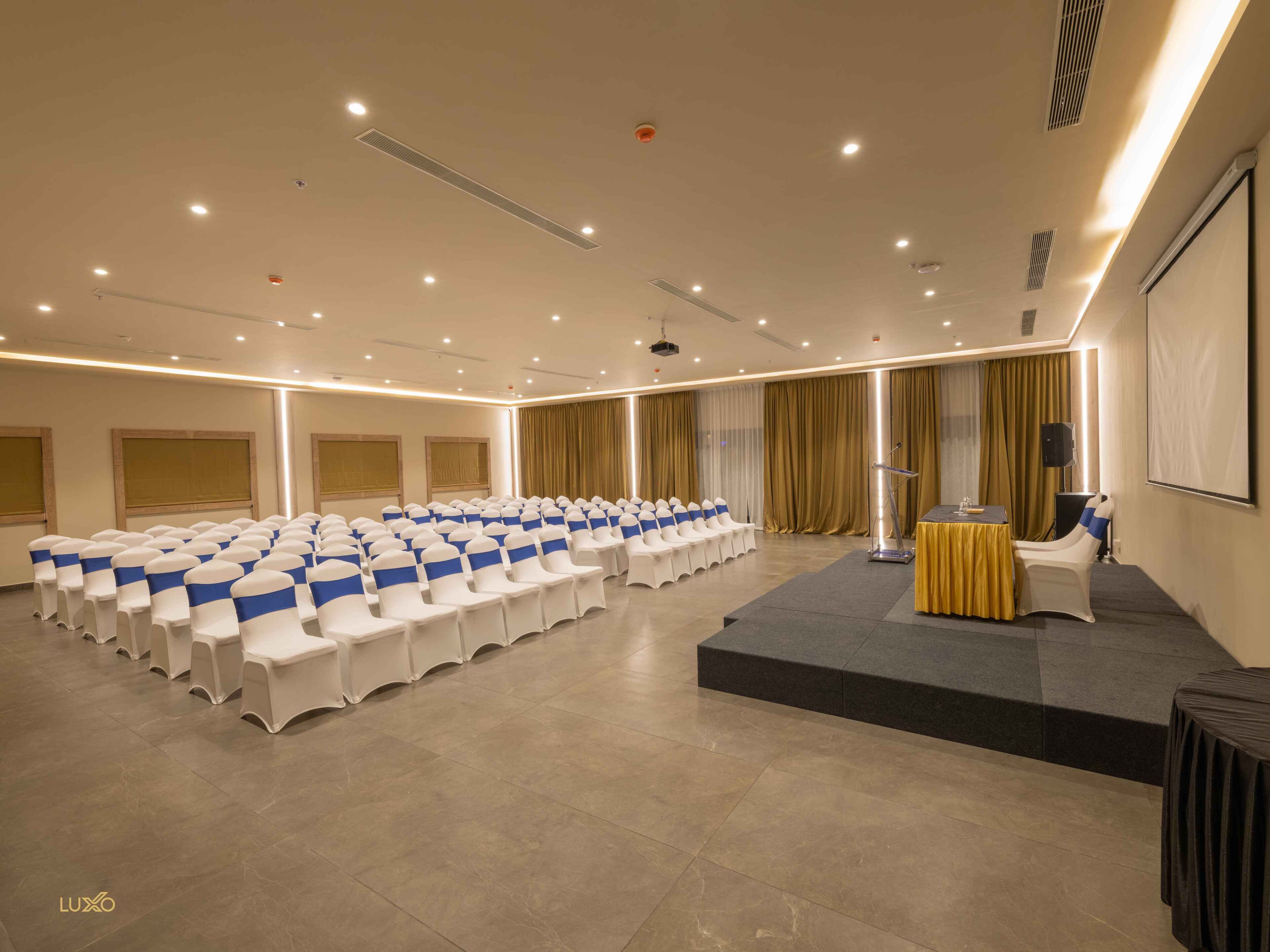 A conference room with rows of white chairs with blue sashes, a podium, and a screen on a raised platform.