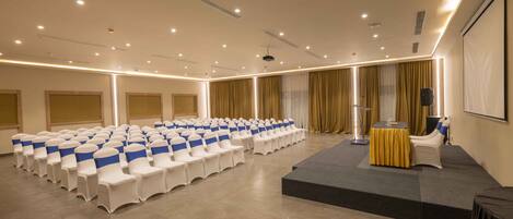 Uma sala de conferências com fileiras de cadeiras brancas com fitas azuis, um pódio e uma tela em uma plataforma elevada.