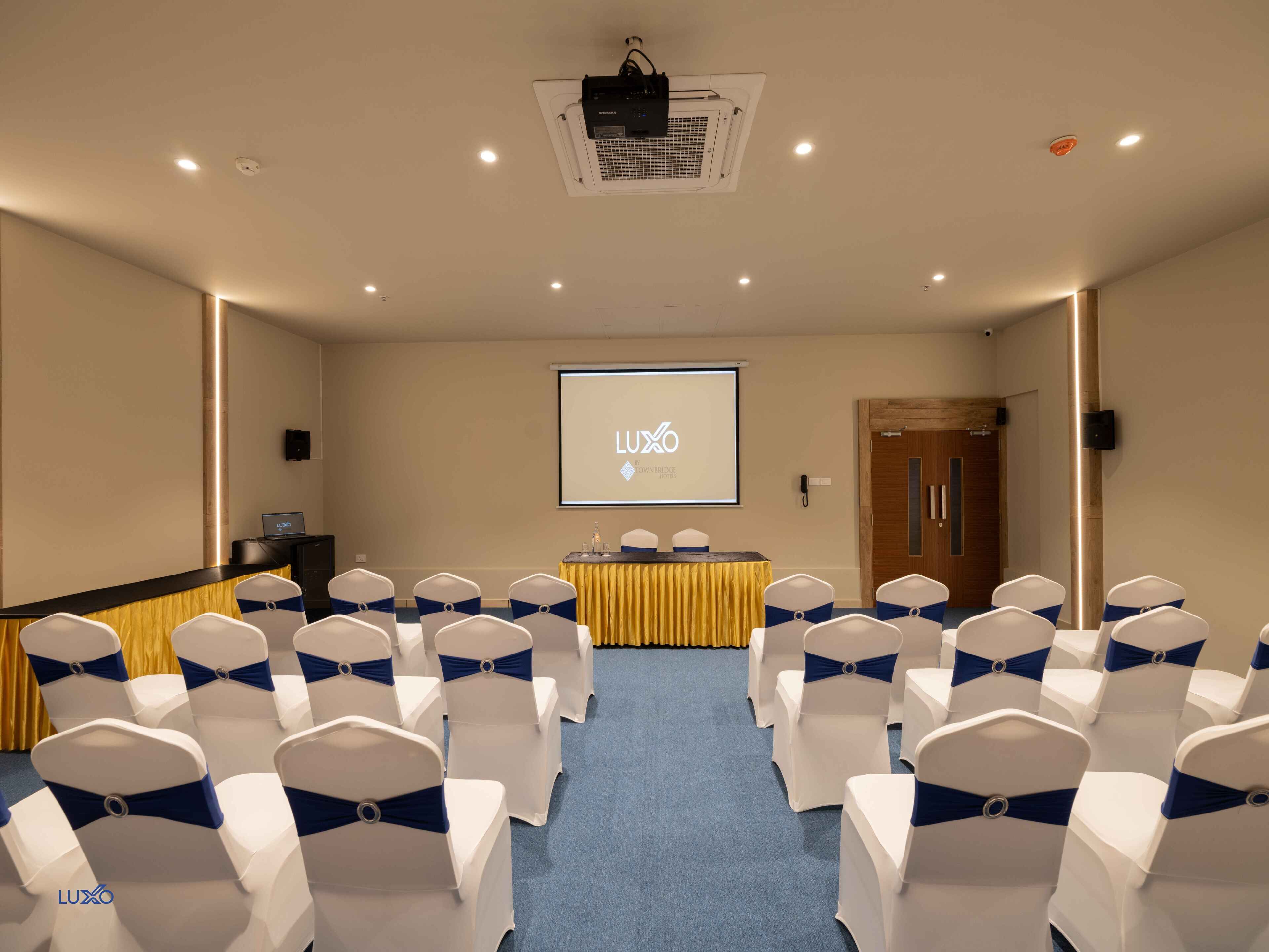 A conference room with rows of white chairs with blue sashes, a podium, a projector screen, and a wooden door.