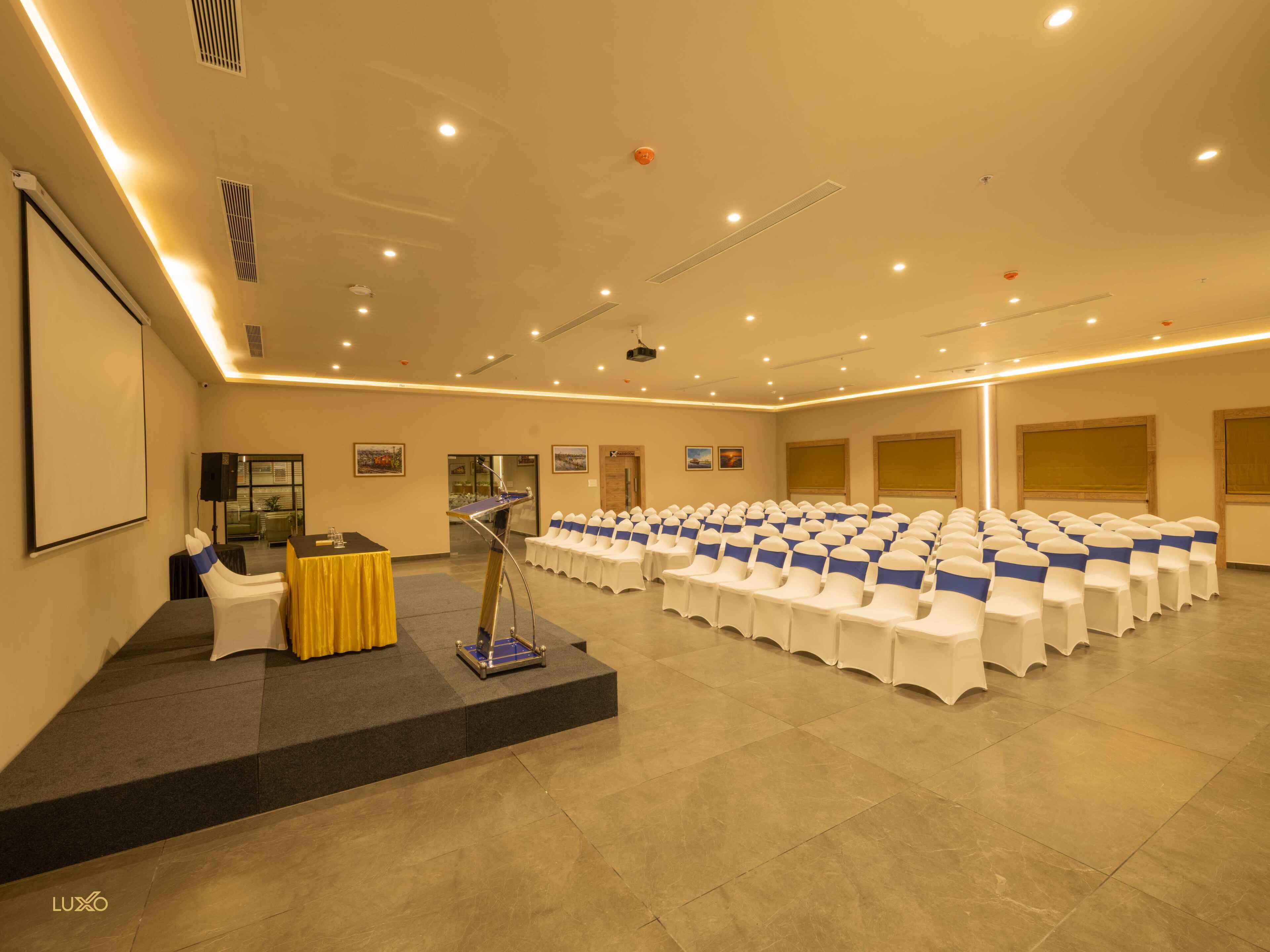 A conference room with rows of white chairs, a podium, and a projector screen.