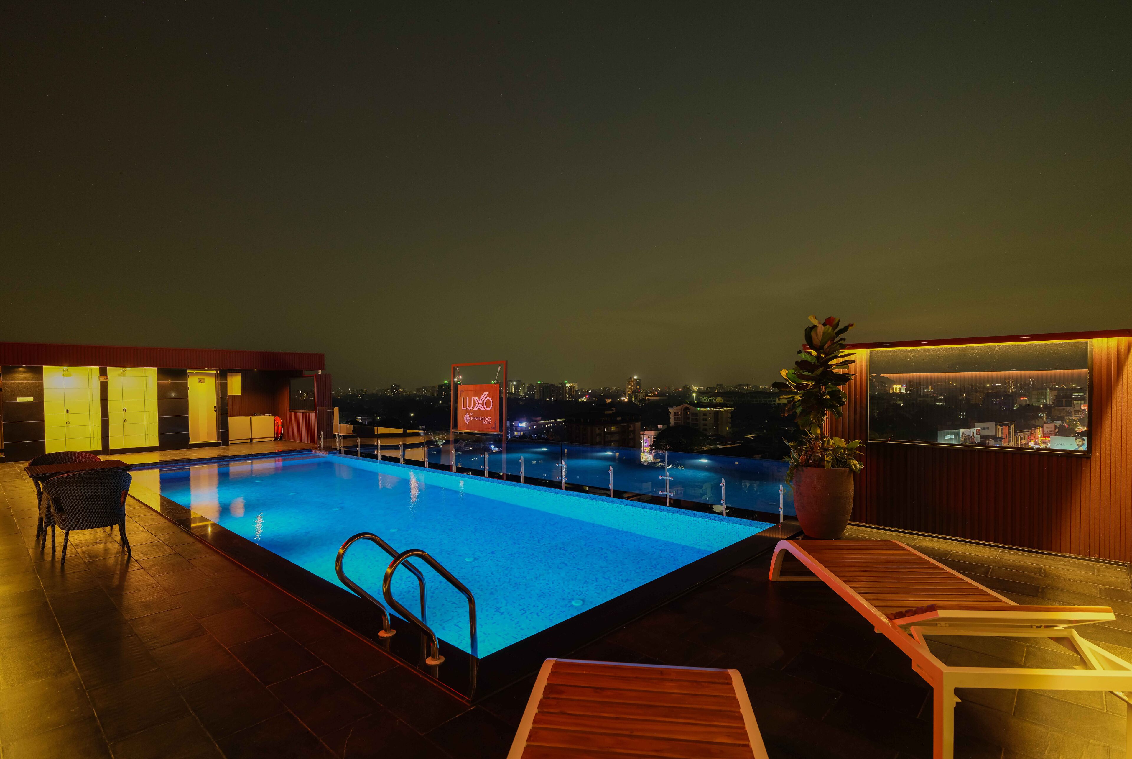 An outdoor swimming pool with a city view at night, a lounge area with chairs, and a sign reading "LUXO".