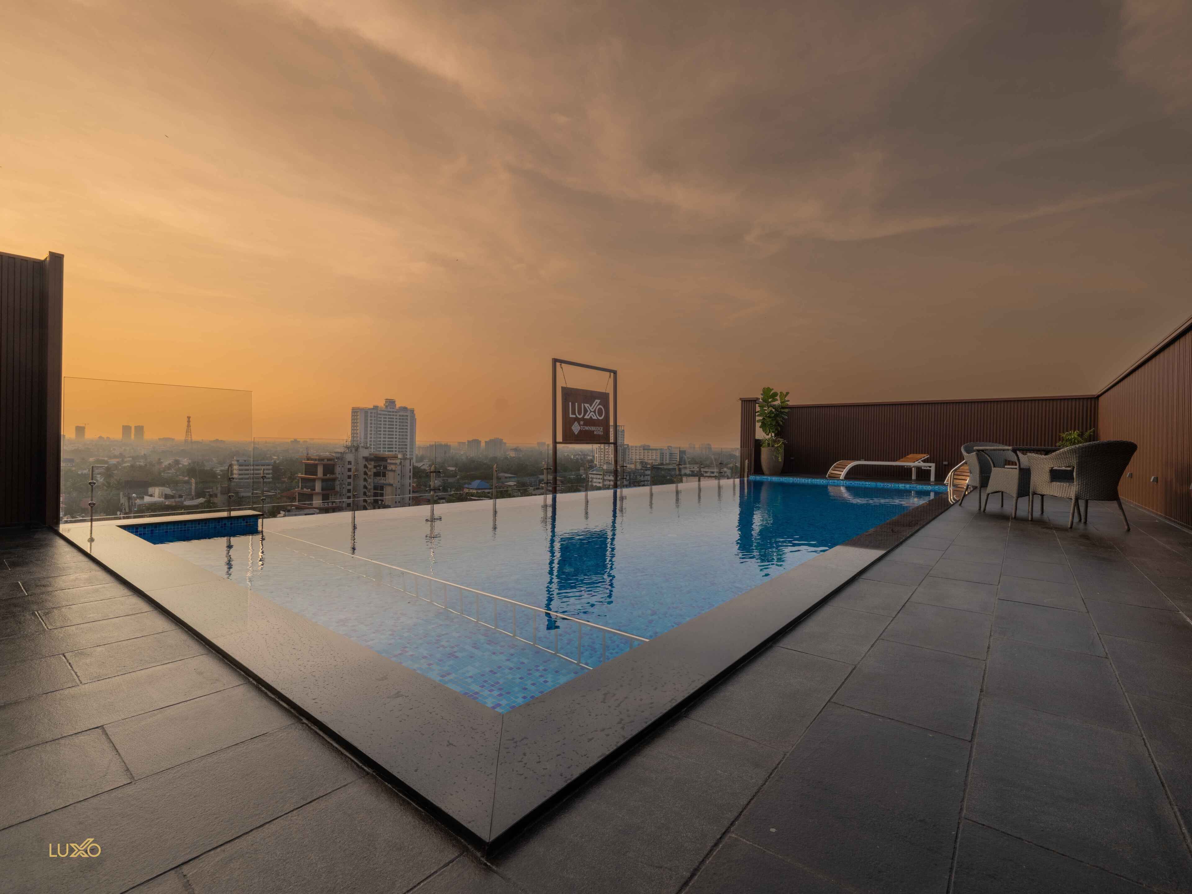 A rooftop infinity pool with a city view, a sign reading "LUXO", and outdoor furniture.