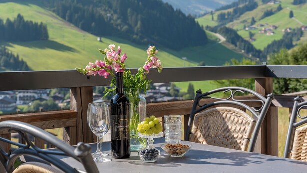 A table set with a bottle of wine, a glass, a vase with flowers, and a bowl of grapes, overlooking a mountainous landscape. - Superb apartment on the slopes with terrace (Hinterglemm)