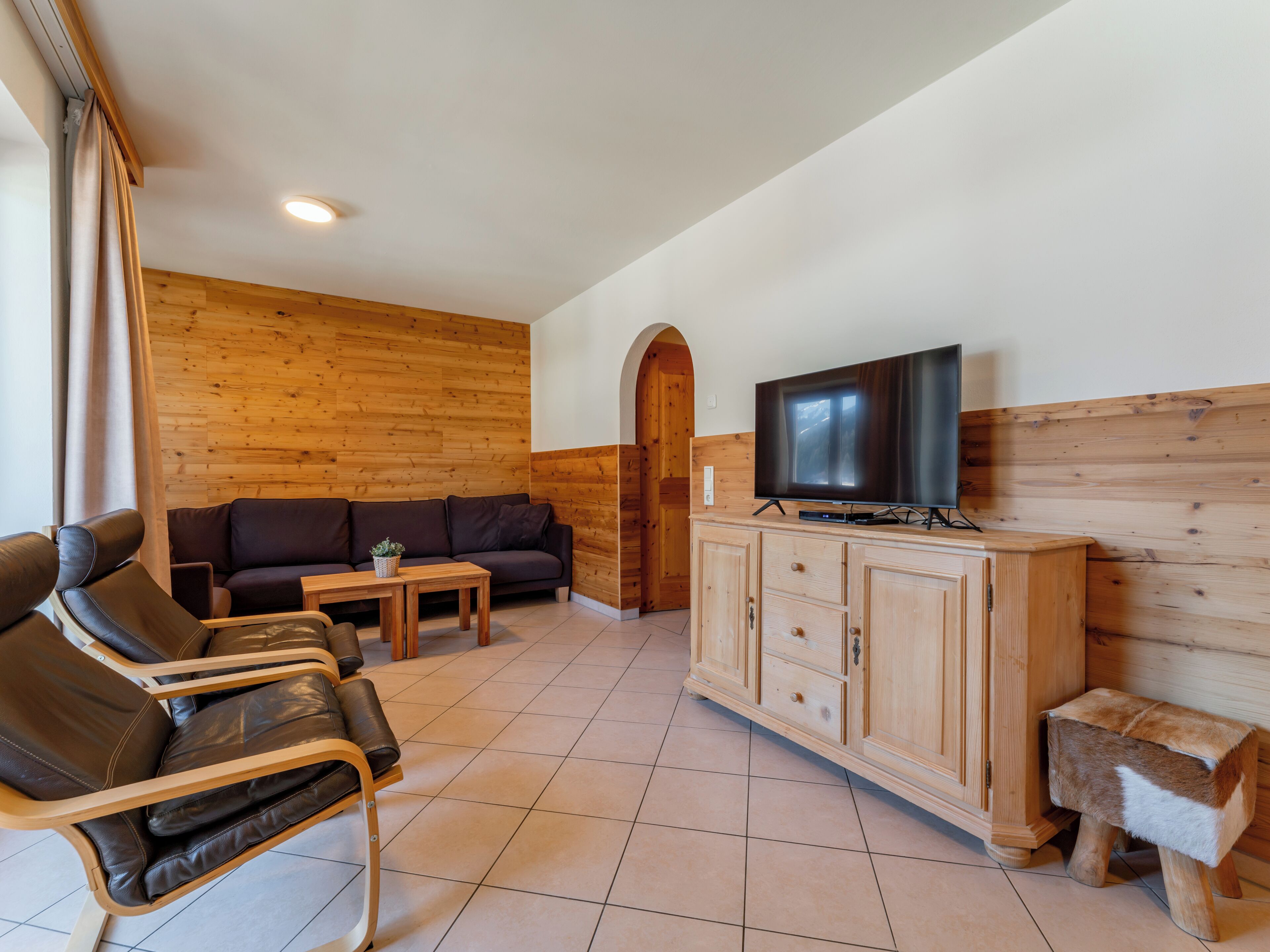 A modern living room with a wooden wall, a leather sofa, a TV on a wooden cabinet, and a small wooden stool.